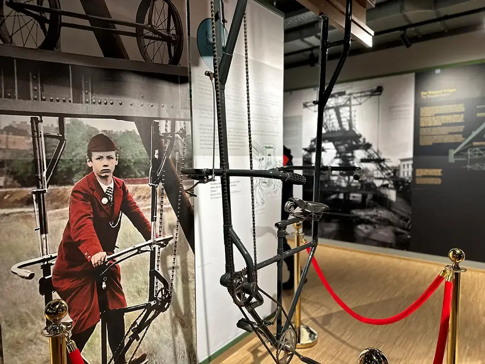 Ein Teil der Ausstellung im Schwebodrom. Ein Junge sitzt auf einem Fahrradähnlichen Gefährt, welches an Schienen aufgehängt ist. 